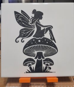 A laser-engraved ceramic tile featuring a fairy perched on a mushroom, engraved from licensed artwork.