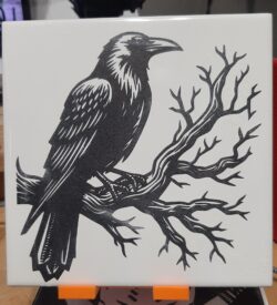A laser-engraved ceramic tile featuring high-contrast crow artwork, engraved from licensed design.